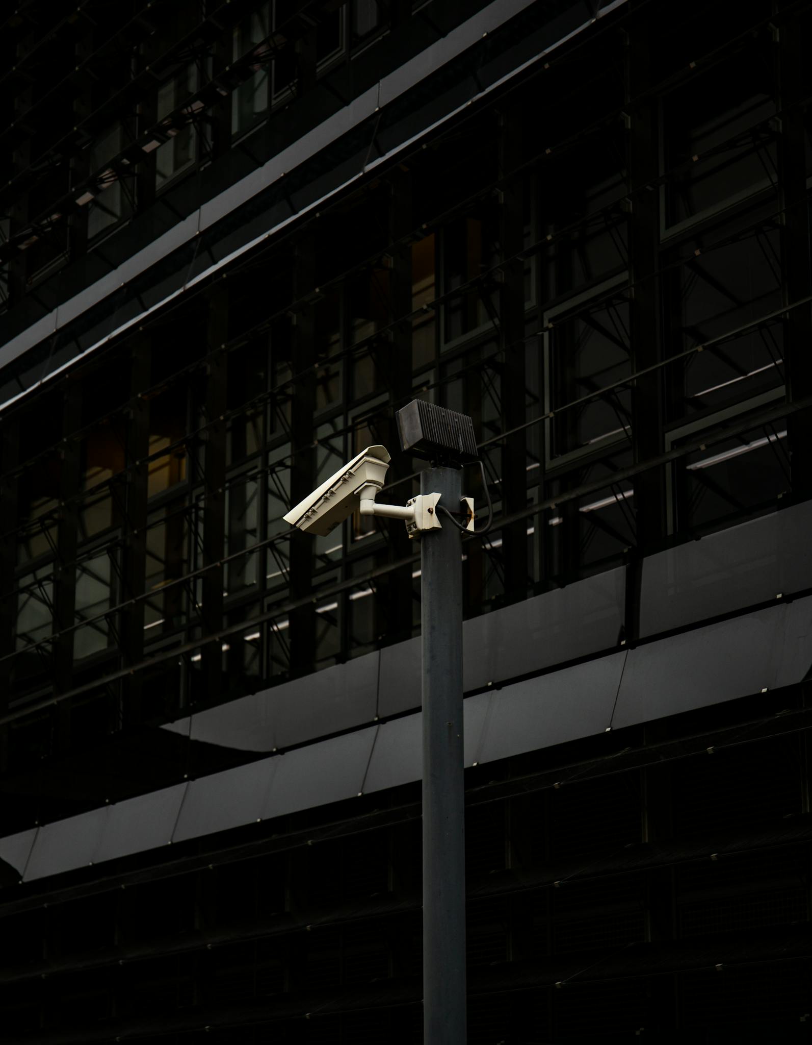 A surveillance camera mounted on a post against a modern urban building in a city setting.